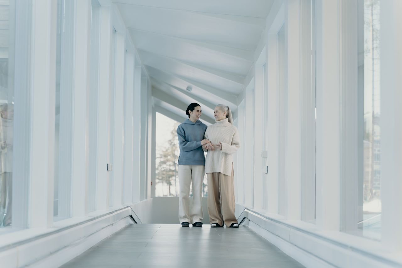 Home Two women walking arm in arm in a bright, modern corridor, exuding joy and companionship.